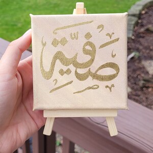 May include: A small canvas painting on an easel with the Arabic word "Subhanallah" written in gold paint.