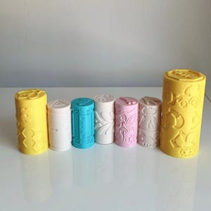 May include: A collection of seven cylindrical plastic rollers in various colours including yellow, white, blue, and pink. Each roller features distinct embossed patterns on its surface, such as flowers, honeycombs, geometric shapes, fairies, and construction vehicles.