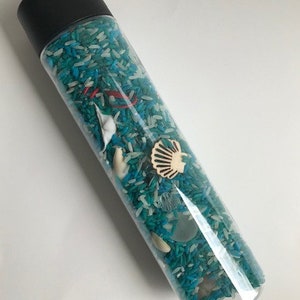 May include: A clear plastic bottle filled with white and blue rice, with various small sea-themed objects, including a seashell, a starfish, and a small piece of driftwood.