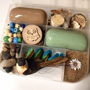 May include: A clear plastic divided container filled with craft supplies. Includes brown and green play dough, wooden beads, small stones, wooden owl cutouts, feathers, and a decorative bird. The container is ideal for crafting and creative projects.