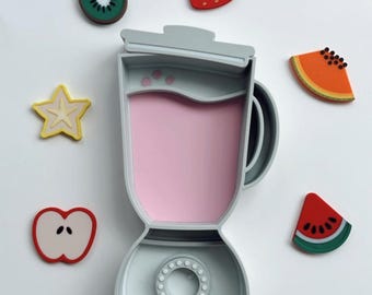 May include: A light gray blender filled with pink liquid, surrounded by colorful fruit-shaped magnets. The magnets include a kiwi, strawberry, starfruit, apple, papaya slice, and watermelon slice.