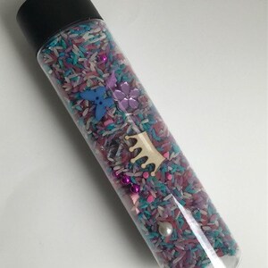 May include: A clear plastic bottle filled with colorful rice, beads, and small plastic shapes. The bottle has a black lid and is decorated with a wooden crown.