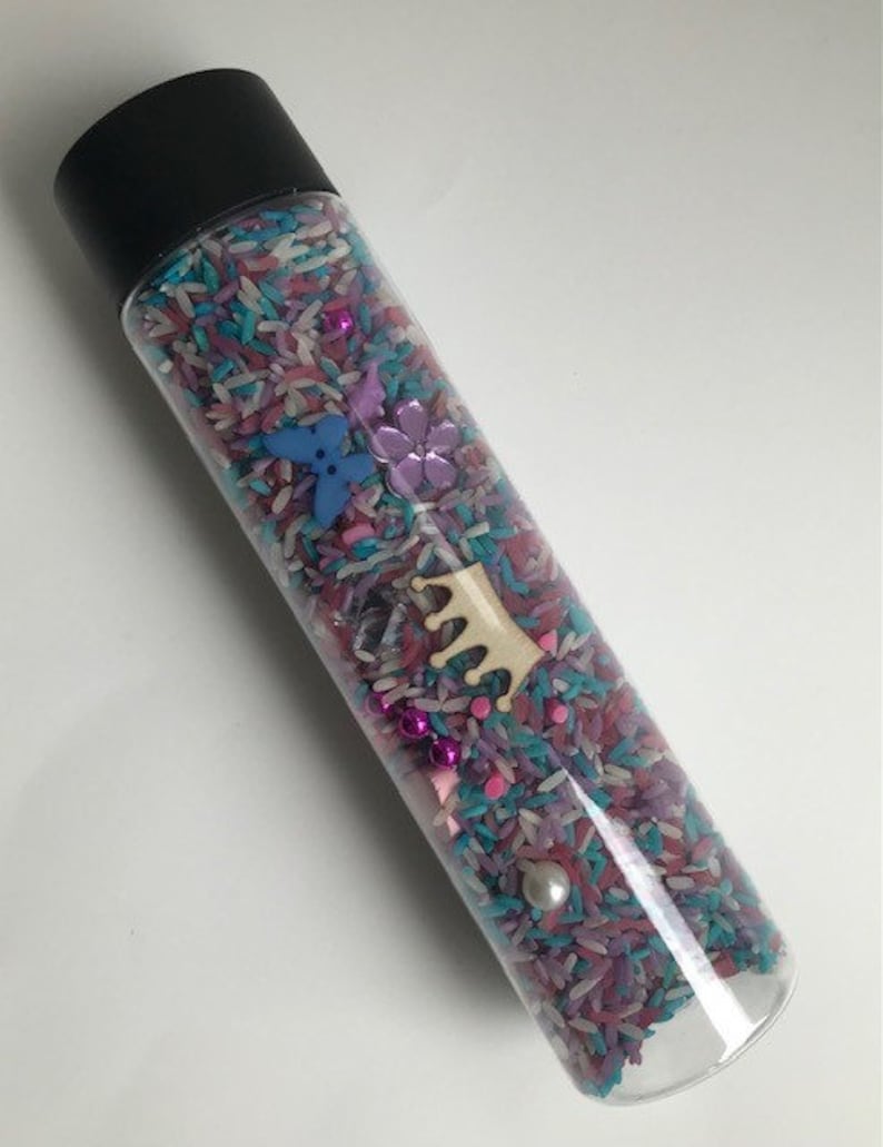 May include: A clear plastic bottle filled with colorful rice, beads, and small plastic shapes. The bottle has a black lid and is decorated with a wooden crown.