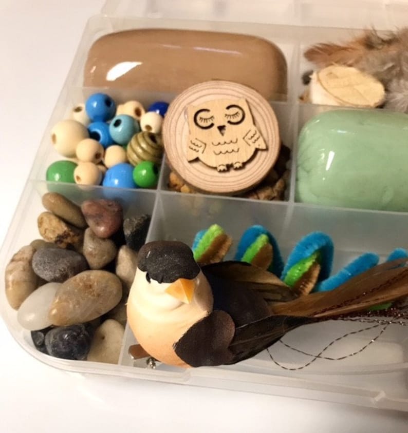 May include: A clear plastic organizer filled with craft supplies. Includes a decorative bird, wooden beads, smooth stones, a wooden owl cutout, and other colorful embellishments. The organizer is divided into several compartments.