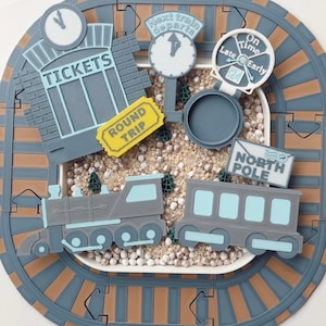 May include: A toy train set featuring a grey and brown track, a grey train, and a passenger carriage. The set includes a ticket booth, clocks, and a sign reading "North Pole". A yellow "Round Trip" ticket is also present.