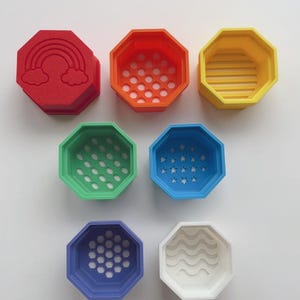 May include: A collection of colorful, octagonal containers in red, orange, yellow, green, blue, purple, and white. Each container features a unique design, including a rainbow, dots, stars, and wavy lines. These containers are likely for storage or decorative purposes.