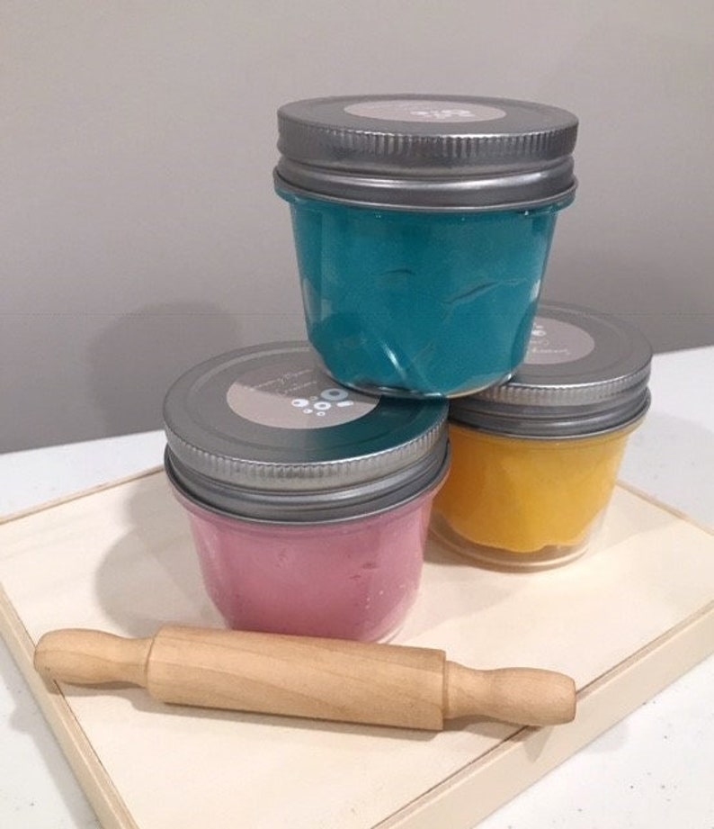 May include: Three glass jars with lids containing different colored play dough. The jars are stacked on top of each other. The play dough is teal, yellow, and pink. A wooden rolling pin is in front of the jars.