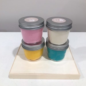 May include: Four small glass jars with silver lids containing different colored slime. The slime colors are pink, white, yellow, and teal.