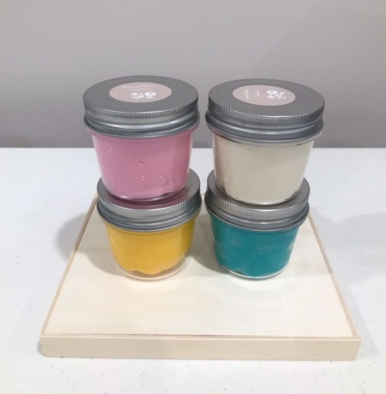 May include: Four small glass jars with silver lids containing different colored slime. The slime colors are pink, white, yellow, and teal.