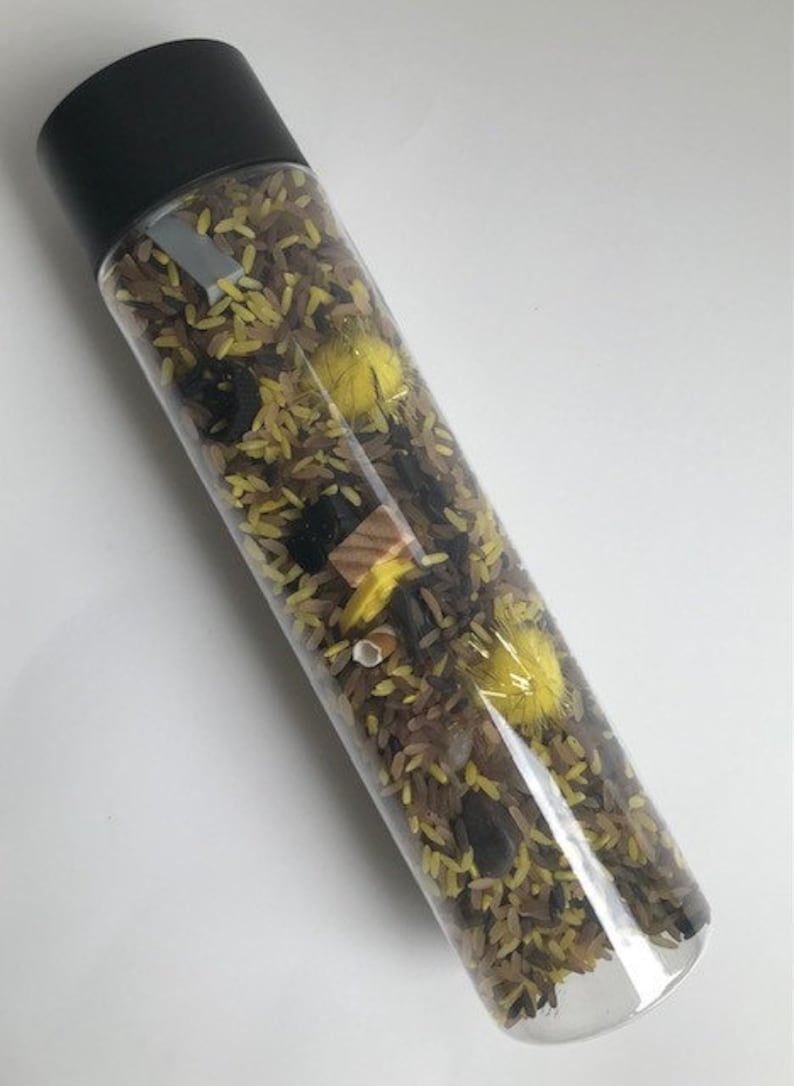 May include: A clear plastic bottle filled with brown and yellow rice, black beans, yellow pom-poms, and a small wooden block.