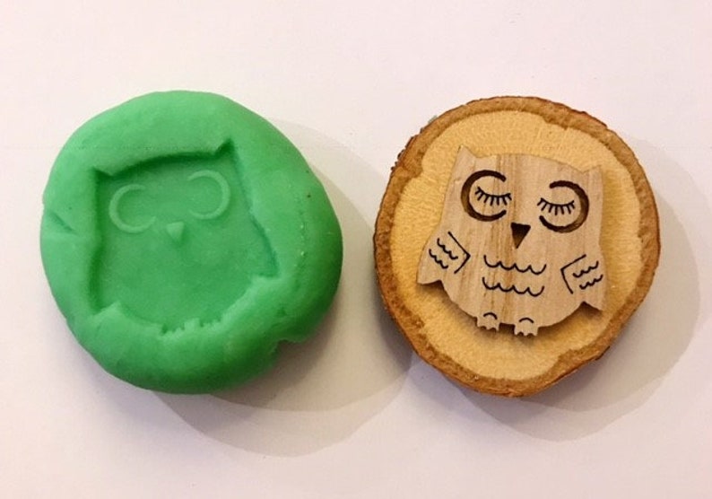 May include: A green, oval-shaped mold with an owl design next to a wooden owl-shaped stamp. The stamp is light brown with a detailed owl design. The owl has closed eyes and wing details.