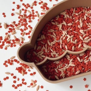 May include: A brown, cloud-shaped container filled with red star-shaped sprinkles and tan, oval-shaped sprinkles. Some sprinkles are scattered on the white surface. The container has a scalloped edge.