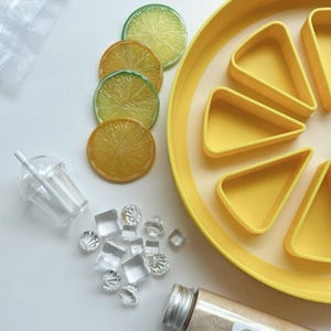 May include: A vibrant composition featuring a yellow lemon-shaped tray with triangular sections, artificial lemon slices, clear ice cubes, a small plastic cup with a straw, and a vial labelled "Lemonade Fizzy Potion".