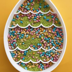 May include: An egg-shaped white tray filled with green rice and colourful beans. The tray has wavy dividers. The beans are various colours, including pink, blue, yellow, and brown. The background is a solid yellow.