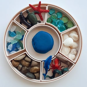 Peut inclure: Un plateau circulaire blanc avec huit compartiments, chacun rempli d'objets sur le thème de l'océan. Les compartiments contiennent de la pâte à modeler bleue, des coquillages, de petits cailloux, des perles de verre, une étoile de mer, une tortue, un dauphin et une pieuvre.