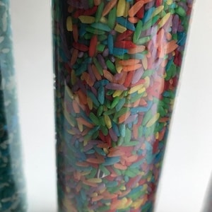 May include: A clear glass cylinder filled with colorful rice. The rice is a mix of red, orange, yellow, green, blue, and purple.