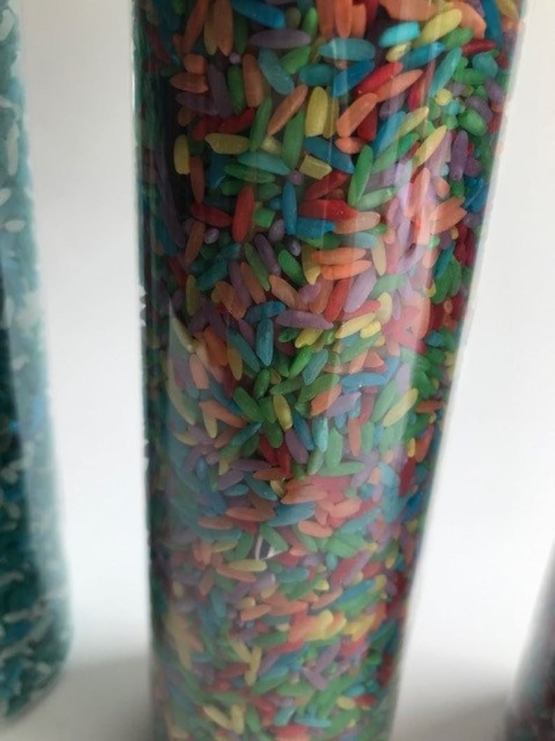 May include: A clear glass cylinder filled with colorful rice. The rice is a mix of red, orange, yellow, green, blue, and purple.