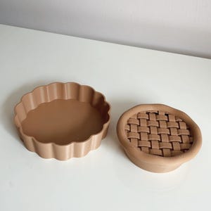 May include: Two tan pie dishes on a white surface. One is a fluted, open pie pan. The other is a round dish with a woven pattern on the top. Both are made of a solid material. The dishes are likely for baking.