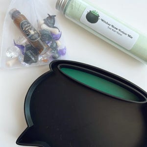 May include: A black cauldron-shaped tray with a green interior, alongside a tube of green powder labeled "Witches Brew Potion Mix" and a small bag containing potion ingredients. The items are arranged on a white surface.