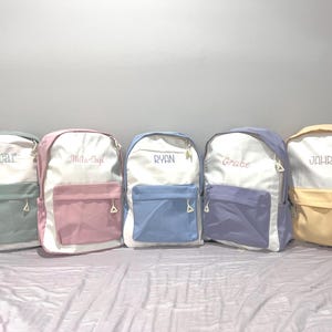 May include: Five pastel-coloured backpacks, each with a name embroidered on the white top section. Colours include green, pink, blue, purple, and yellow. The front pocket and side panels match the backpack's colour scheme.