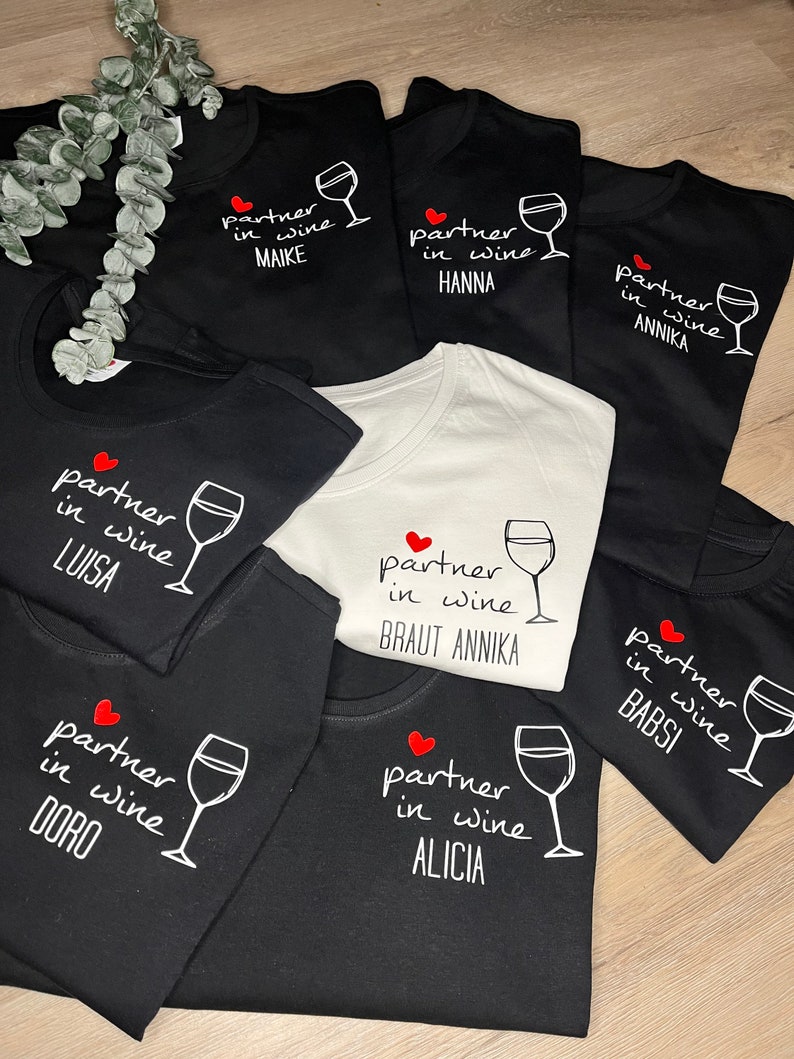 Pu&ograve; includere: Sette magliette nere con testo bianco e un design a forma di cuore rosso. Il testo sulle magliette recita "partner in wine" seguito da un nome. Una maglietta ha il testo "partner in wine BRAUT ANNIKA".