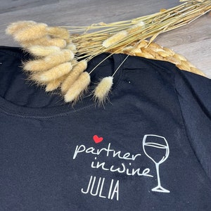 Pu&ograve; includere: Maglietta nera con testo bianco che recita "partner in wine JULIA" e un cuore rosso e un'immagine di un bicchiere di vino bianco.