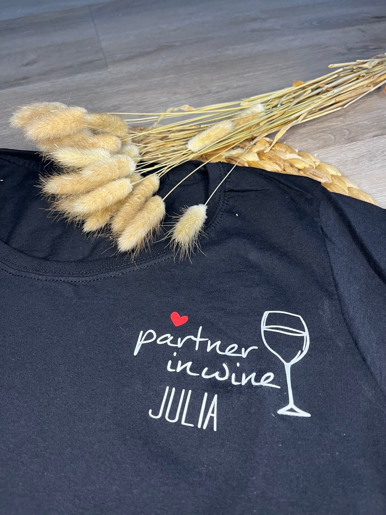 Pu&ograve; includere: Maglietta nera con testo bianco che recita "partner in wine JULIA" e un cuore rosso e un'immagine di un bicchiere di vino bianco.