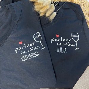 Pu&ograve; includere: Due magliette nere con una grafica di bicchiere di vino bianco e il testo "partner in wine" con un cuore rosso. Una maglietta dice "Katharina" e l'altra dice "Julia".