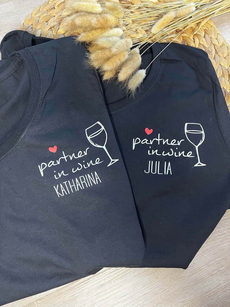 Pu&ograve; includere: Due magliette nere con una grafica di bicchiere di vino bianco e il testo "partner in wine" con un cuore rosso. Una maglietta dice "Katharina" e l'altra dice "Julia".