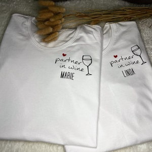 Pu&ograve; includere: Due magliette bianche con il testo "partner in wine" e un'immagine di un bicchiere di vino. Una maglietta dice "Marie" e l'altra dice "Linda".