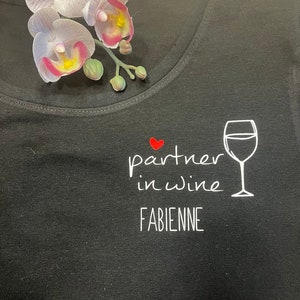 Pu&ograve; includere: Maglietta nera con testo bianco che recita "partner in wine" con un cuore rosso e un'immagine di un bicchiere di vino bianco. Il nome "FABIENNE" &egrave; stampato sotto il testo.