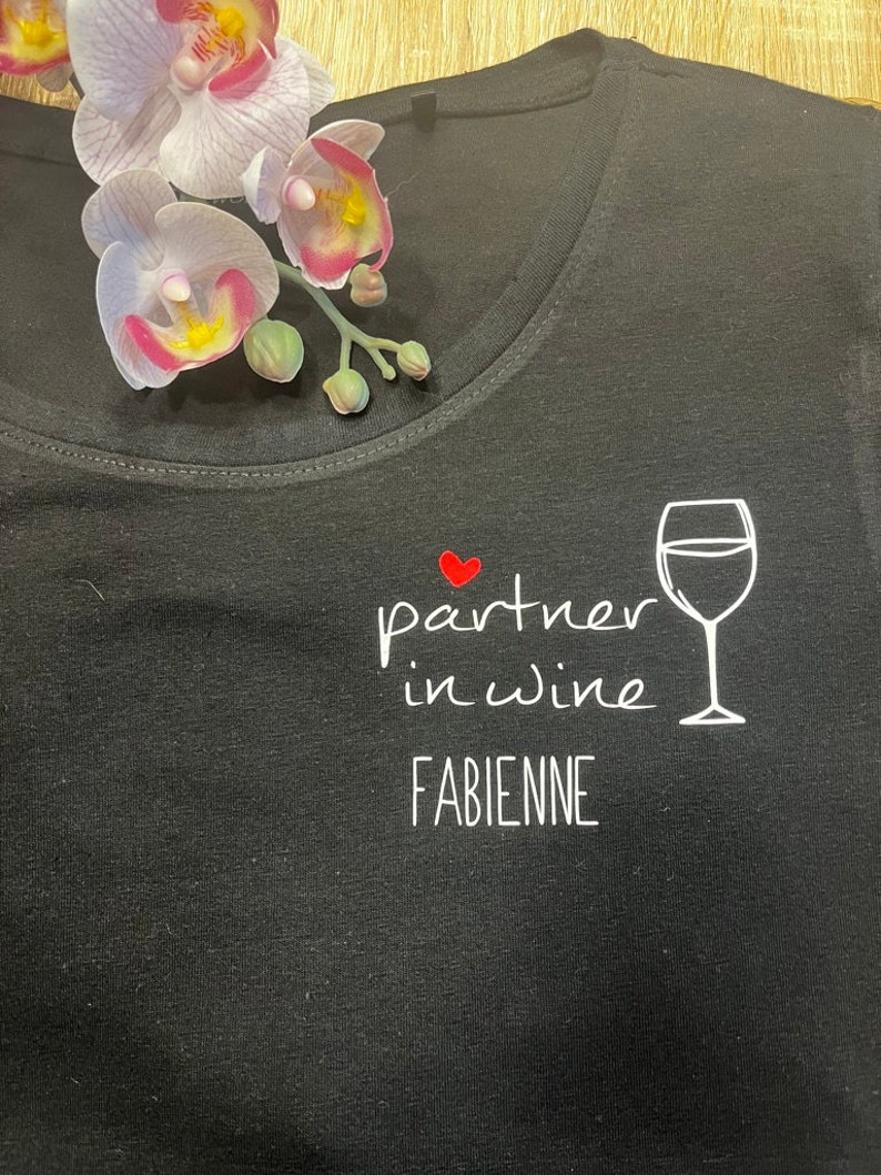Pu&ograve; includere: Maglietta nera con testo bianco che recita "partner in wine" con un cuore rosso e un'immagine di un bicchiere di vino bianco. Il nome "FABIENNE" &egrave; stampato sotto il testo.