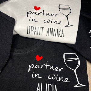 Pu&ograve; includere: Due magliette bianche e nere con un cuore rosso e un bicchiere di vino. Il testo sulla maglietta bianca dice "partner in wine Braut Annika". Il testo sulla maglietta nera dice "partner in wine Alicia".