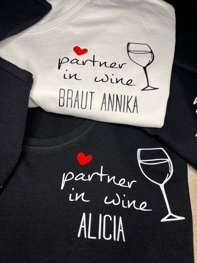 Pu&ograve; includere: Due magliette bianche e nere con un cuore rosso e un bicchiere di vino. Il testo sulla maglietta bianca dice "partner in wine Braut Annika". Il testo sulla maglietta nera dice "partner in wine Alicia".