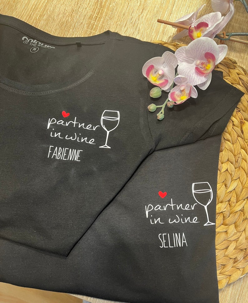 Pu&ograve; includere: Due magliette nere con il testo "partner in wine" e un'immagine di un bicchiere di vino. La prima maglietta ha il nome "FABIENNE" stampato sotto l'immagine. La seconda maglietta ha il nome "SELINA" stampato sotto l'immagine.