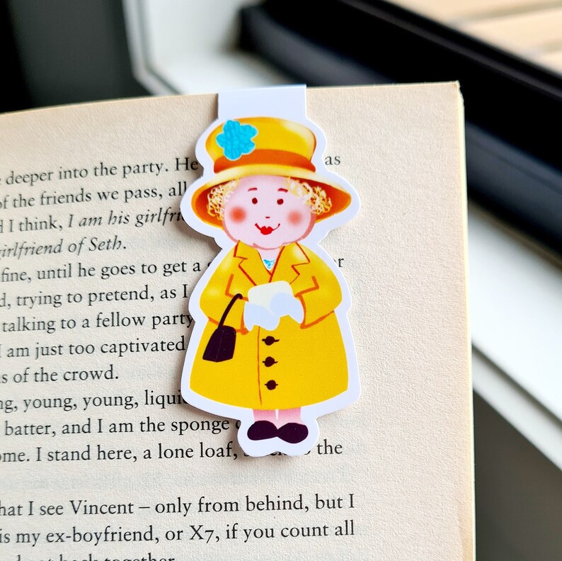 May include: A yellow bookmark with a cartoon image of a woman wearing a yellow coat, a white hat with a blue flower, and black shoes. The woman is holding a white envelope in her right hand and a black purse in her left hand.