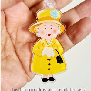May include: A yellow plastic keychain featuring a cartoon illustration of a woman wearing a yellow dress, a yellow hat with a blue flower, and a black purse. The woman has blonde hair and a smiling face.