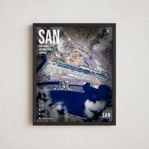 May include: A framed aerial map print of San Diego International Airport, labeled SAN. The image features a satellite view of runways, terminals, and nearby water, overlaid with white text, airport codes, and coordinates against a dark background.