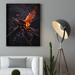 Könnte beinhalten: Ein gerahmtes Foto eines Lagerfeuers mit orangefarbenen Flammen und brennendem Holz vor einem dunklen Hintergrund. Eine weiße Couch mit einem grauen Kissen und einer kleinen grünen Topfpflanze steht auf einem Holzboden. Eine hohe schwarze Stehlampe mit einem weißen Lampenschirm steht in der Ecke des Raumes.