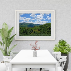 以下が含まれることがあります： 白い壁に白い額縁に入った緑の丘陵地帯の絵が飾られています。青い空に白い雲が浮かんでいます。絵の下には白いテーブルと白い椅子が置かれています。