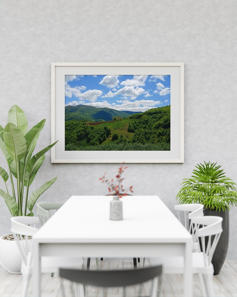 以下が含まれることがあります： 白い壁に白い額縁に入った緑の丘陵地帯の絵が飾られています。青い空に白い雲が浮かんでいます。絵の下には白いテーブルと白い椅子が置かれています。