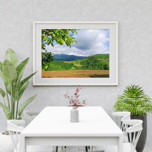 K&ouml;nnte beinhalten: Ein wei&szlig; gerahmtes Bild einer gr&uuml;nen H&uuml;gellandschaft mit bew&ouml;lktem Himmel. Das Bild h&auml;ngt an einer wei&szlig;en Wand &uuml;ber einem wei&szlig;en Tisch mit wei&szlig;en St&uuml;hlen.