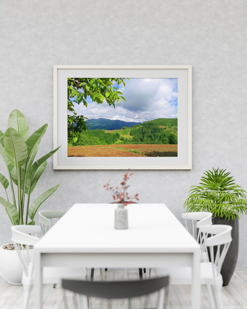 K&ouml;nnte beinhalten: Ein wei&szlig; gerahmtes Bild einer gr&uuml;nen H&uuml;gellandschaft mit bew&ouml;lktem Himmel. Das Bild h&auml;ngt an einer wei&szlig;en Wand &uuml;ber einem wei&szlig;en Tisch mit wei&szlig;en St&uuml;hlen.