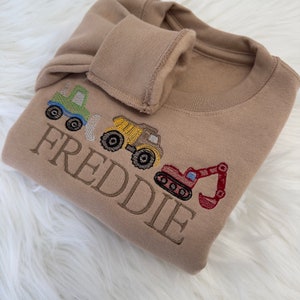 May include: A tan sweatshirt with embroidered construction vehicles and the name "FREDDIE" in brown thread.