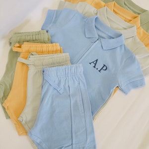 May include: A set of clothing items in various shades of blue, green, and yellow. The set includes a light blue polo shirt with a zipper and the initials "A.P." embroidered on the chest, a pair of light blue shorts, and several pairs of shorts in different shades of green and yellow.