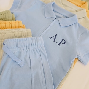 May include: A light blue polo shirt with a zipper and the initials "A.P." embroidered on the chest. The shirt is layered on top of a stack of light blue, beige, and yellow pants.