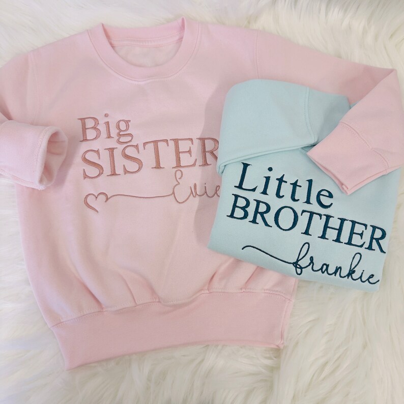 May include: Two pink and light blue sweatshirts with embroidered text. The pink sweatshirt says "Big Sister Evie" with a heart symbol. The light blue sweatshirt says "Little Brother Frankie".