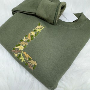 May include: A green sweatshirt featuring an embroidered design of dinosaurs in various shades of green, brown, and yellow.