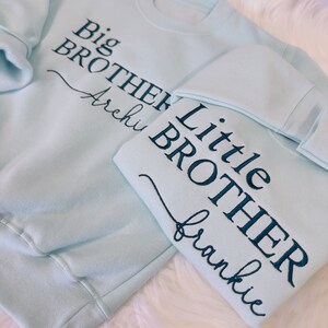 May include: Two light blue sweatshirts with embroidered text. The larger sweatshirt says "Big Brother Archi" and the smaller sweatshirt says "Little Brother Frankie".