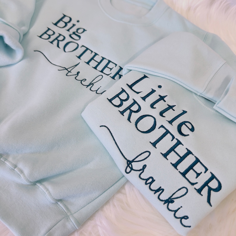 May include: Two light blue sweatshirts with embroidered text. The larger sweatshirt says "Big Brother Archi" and the smaller sweatshirt says "Little Brother Frankie".